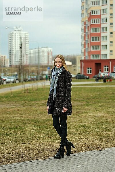 Fröhliches Mädchen in Jeans und Daunenjacke geht die Straße in einem Wohngebiet mit hohen Häusern entlang