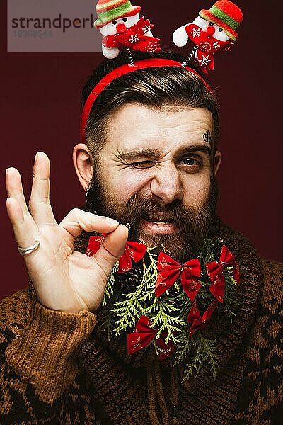 Lustiger bärtiger Mann in einem Neujahrsbild als Weihnachtsmann mit Dekorationen auf seinem Bart. Fest der Weihnacht. Fotos im Studio aufgenommen