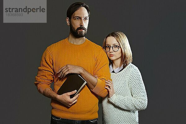 Mann mit Bart im Pullover hält ein Buch in der Hand und ein Mädchen mit Brille hält seinen Arm. Junges ernstes Paar