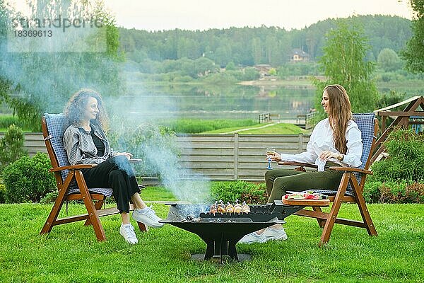 Zwei junge Frauen im Hinterhof  die auf Stühlen sitzen und sich in der Nähe des Grills mit Gemüse unterhalten