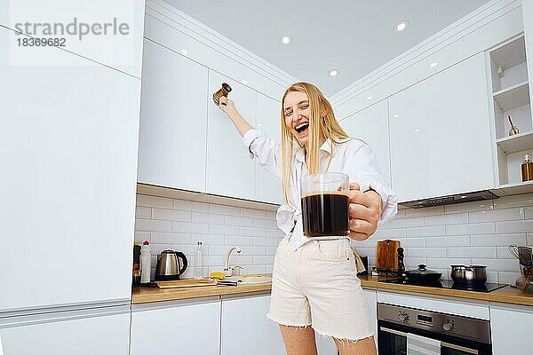 Fröhliche Frau tanzend und schreiend mit einer Tasse Kaffee in der Hand in der Küche