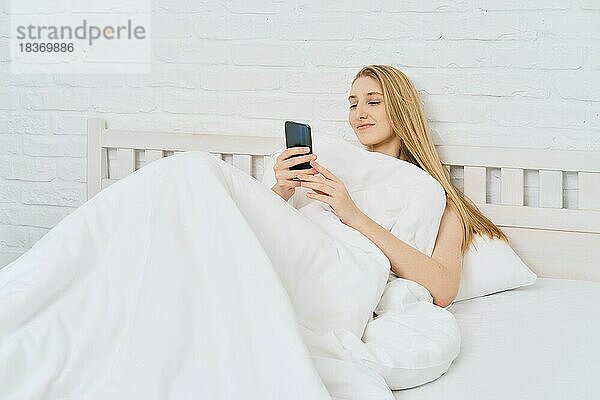 Junge Frau wacht auf und schreibt im Bett eine SMS auf dem Mobiltelefon