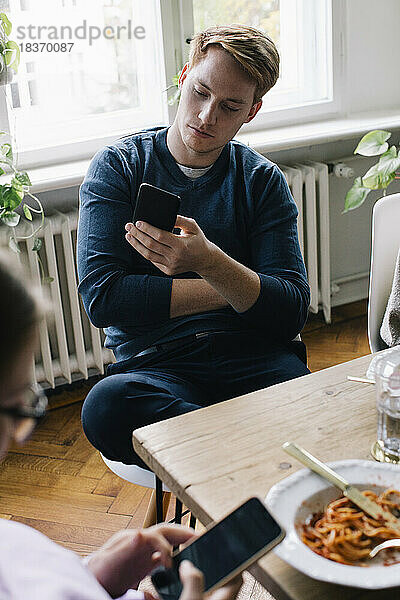 Junger Mann mit Smartphone am Esstisch sitzend in einem Haus