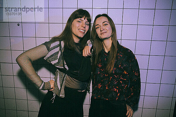 Porträt einer lächelnden Transgender-Frau mit einer Freundin  die in einem Nachtclub an einer weißen Wand steht