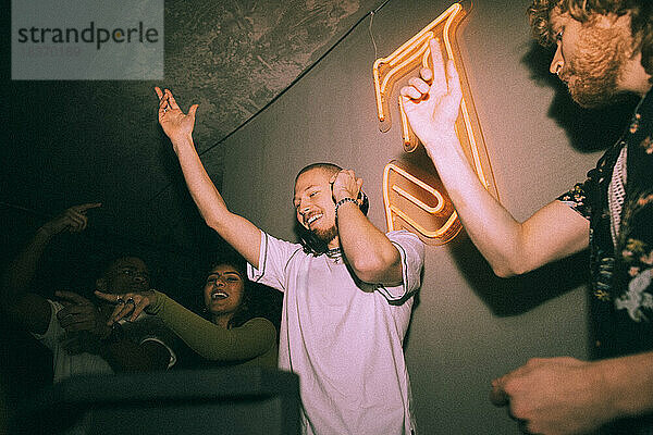 Fröhlicher junger DJ gestikuliert inmitten von Freunden  die in einem beleuchteten Nachtclub tanzen
