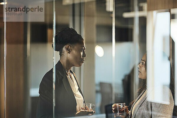 Seitenansicht von Geschäftsfrauen  die im Büro miteinander diskutieren  gesehen durch Glas