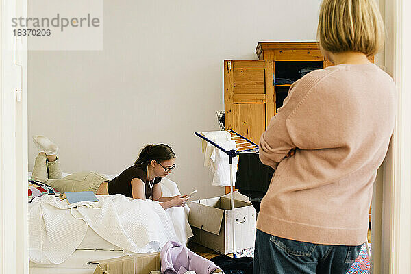 Rückansicht eines Elternteils  der seine Tochter beim Benutzen eines Smartphones beobachtet  die zu Hause auf dem Bett liegt