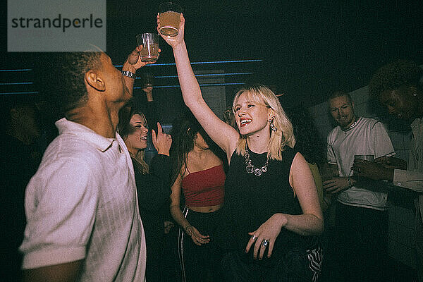 Glückliche junge Frau und Mann stoßen mit Getränken an  während sie in einem Nachtclub mit Freunden tanzen