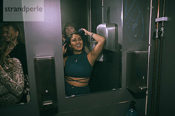 Glückliche junge Frau  die sich mit Freunden amüsiert  während sie ihr Spiegelbild im Badezimmer eines Nachtclubs betrachtet