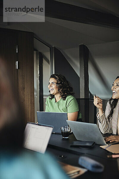 Reife Geschäftskollegen mit Laptops  die lachend an einem Schreibtisch in einer Bürobesprechung sitzen