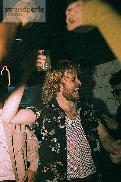 Fröhlicher junger Mann mit Getränk in der Hand tanzt mit Freunden in einem Nachtclub