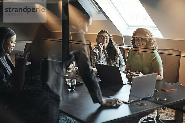 Unternehmerinnen diskutieren über einen Laptop  den sie bei einer Sitzung im Büro durch Glas sehen
