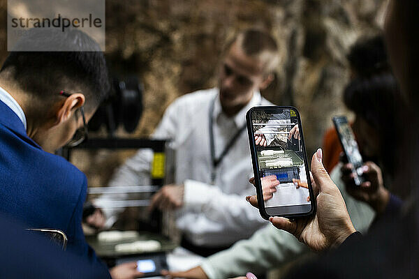 Geschäftsmann stellt 3D-Drucker aus  während Kollegen im Kongresszentrum mit Smartphones filmen