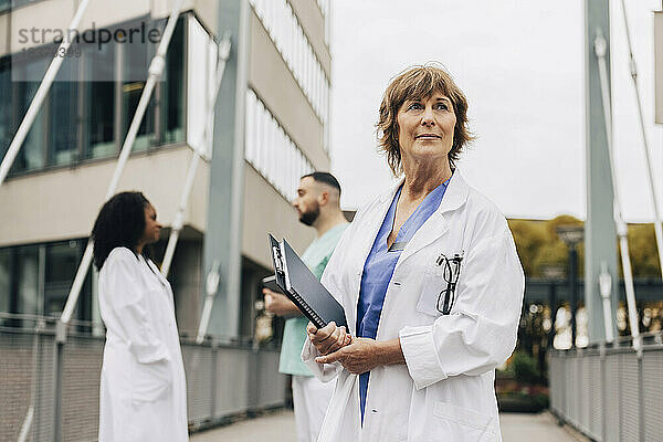 Reife Ärztin schaut weg  während sie ein Klemmbrett und ein Buch außerhalb des Krankenhauses hält