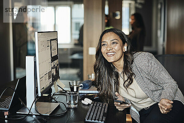 Glückliche Geschäftsfrau mit drahtlosen In-Ear-Kopfhörern  die am Schreibtisch im Büro sitzt