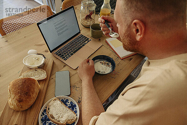 Hoher Blickwinkel eines Mannes  der frühstückt  während er den Laptop auf dem Tisch zu Hause beobachtet
