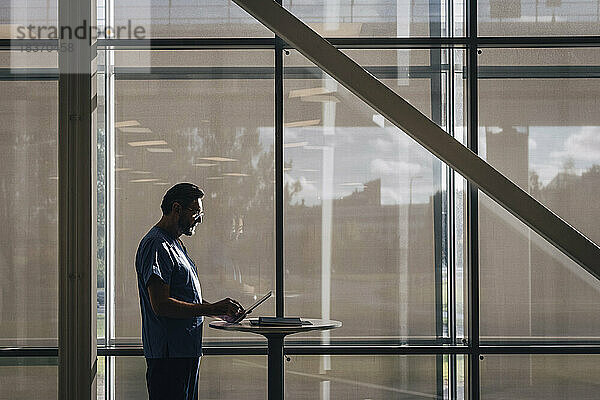 Seitenansicht eines männlichen Arztes  der einen Tablet-PC benutzt  während er an einem Tisch im Krankenhaus steht
