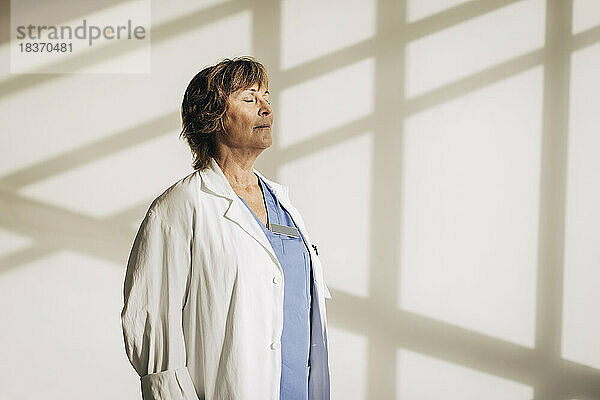 Ältere Ärztin steht mit geschlossenen Augen vor einer Wand mit Schatten