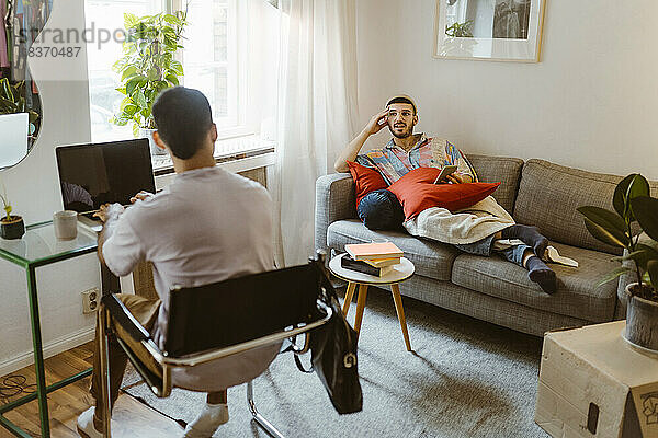 Mann auf dem Sofa im Gespräch mit seinem Freund  der zu Hause am Laptop sitzt