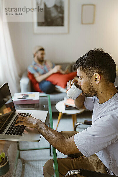 Seitenansicht eines Mannes  der zu Hause Kaffee trinkt und einen Laptop benutzt