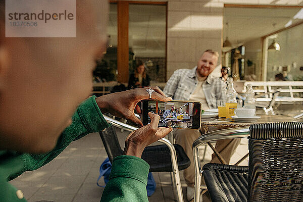 Junger Mann fotografiert männlichen Freund mit seinem Smartphone in einem Straßencafé