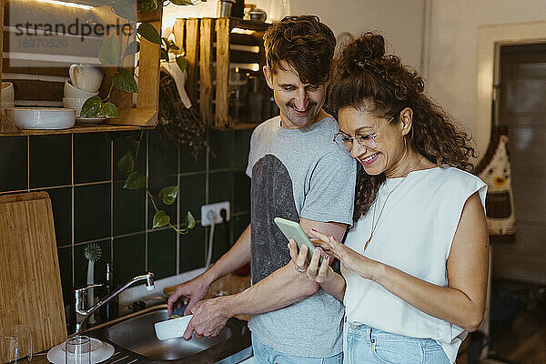 Ein lächelnder Mann wäscht ab und schaut dabei auf das Smartphone seiner Freundin in der Küche