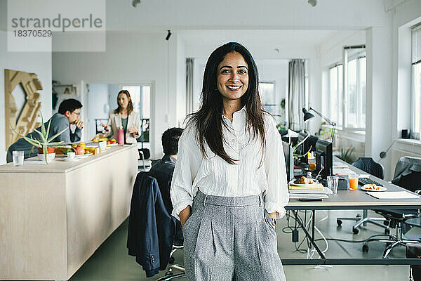 Porträt einer zuversichtlich lächelnden Geschäftsfrau  die mit den Händen in den Taschen im Büro steht
