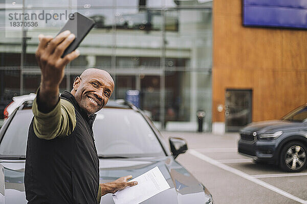 Glücklicher Mann  der nach dem Kauf eines neuen Autos ein Selfie mit seinem Smartphone macht