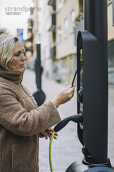 Ältere Frau  die an einer Ladestation für Elektrofahrzeuge mit einer digitalen Geldbörse bezahlt