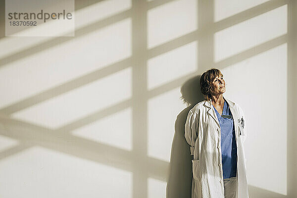 Weiblicher Arzt im Laborkittel träumt  während er gegen eine Wand mit Sonnenlicht steht