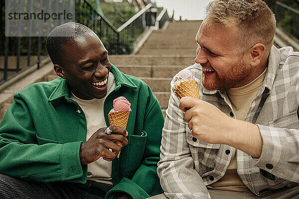 Glückliche multirassische männliche Freunde  die auf einer Treppe sitzend ein Eis genießen