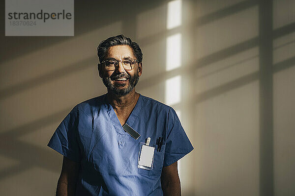 Porträt eines glücklichen männlichen Arztes  der in einem Krankenhaus an der Wand steht