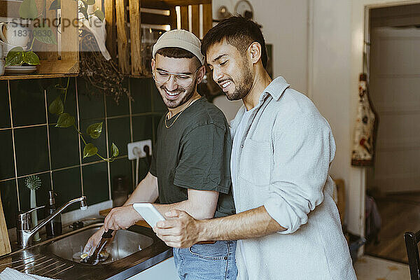 Lächelnder Mann  der sein Smartphone mit seinem Freund teilt  der in der Küche zu Hause abwäscht