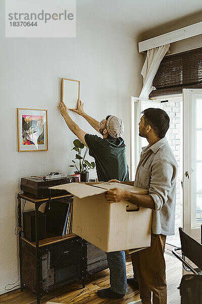 Seitenansicht eines Mannes mit Karton  der bei seinem Freund steht  der zu Hause einen Rahmen an die Wand montiert