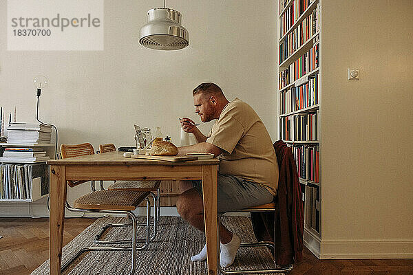 Seitenansicht eines übergewichtigen Mannes  der frühstückt  während er am heimischen Tisch den Laptop betrachtet
