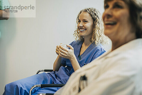 Glückliche Krankenschwester  die eine Kaffeetasse hält  während sie auf einem Stuhl neben einem Kollegen sitzt