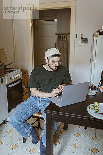 Mann mit Laptop am Esstisch in der Küche zu Hause sitzend