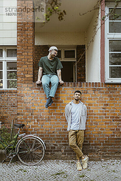 Mann sieht seinen Freund an  der sich mit dem Fahrrad vor dem Haus an eine Mauer lehnt