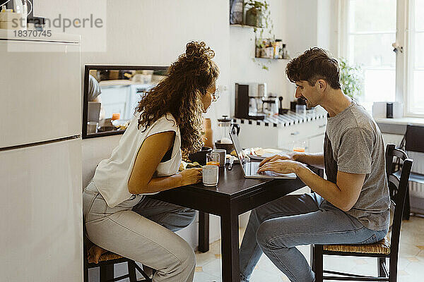 Mann spricht mit Frau  während er einen Laptop auf dem Esstisch zu Hause benutzt