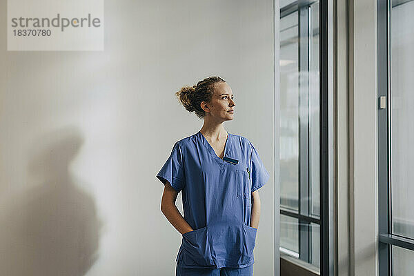 Nachdenkliche junge Krankenschwester steht mit den Händen in den Taschen an der Wand eines Krankenhauses