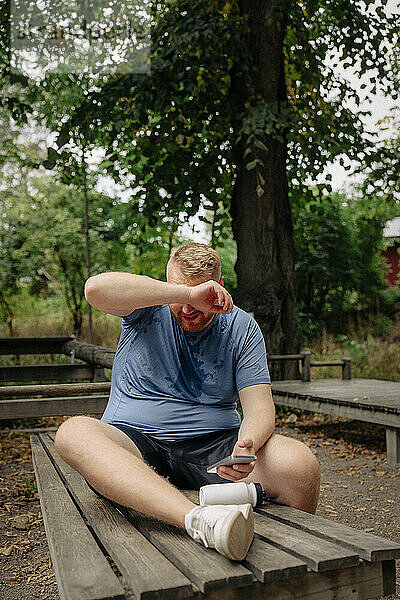 Übergewichtiger Mann mit Smartphone wischt sich den Schweiß ab  während er auf einer Bank im Park sitzt