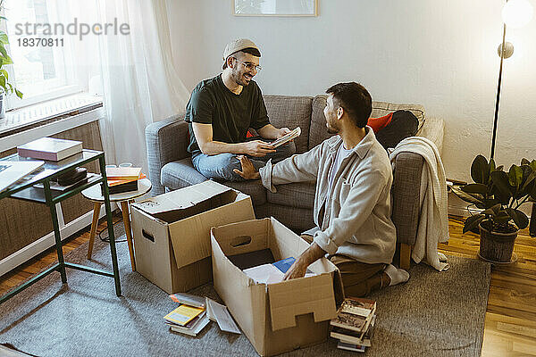 Lächelnder Mann  der auf dem Sofa sitzt und sich mit seinem Freund unterhält  während er zu Hause einen Karton auspackt