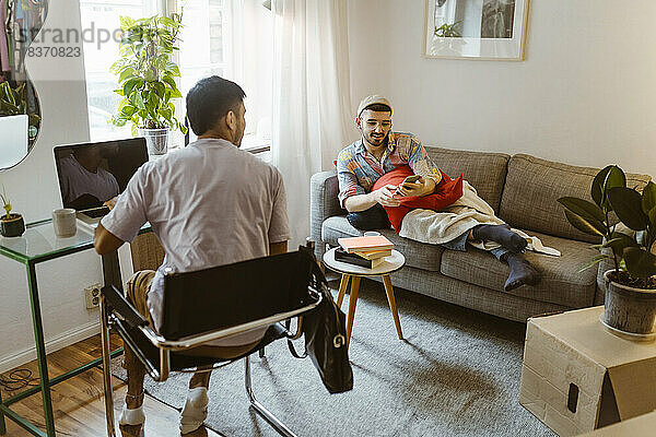Mann sitzt auf einem Stuhl und spricht mit seinem Freund  der ein Smartphone im Wohnzimmer benutzt