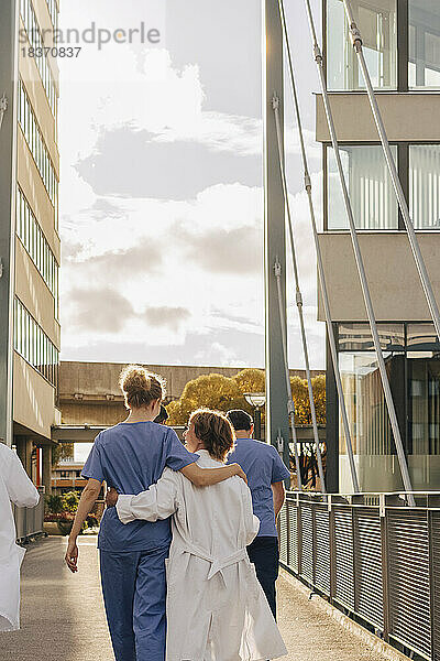 Rückansicht von Mitarbeitern des Gesundheitswesens  die sich gegenseitig umarmen und bei Sonnenuntergang in Richtung Krankenhaus gehen