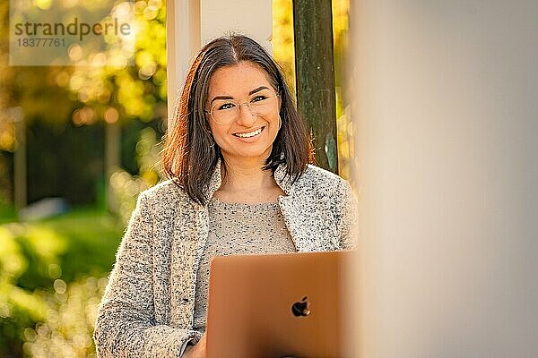 Hübsche Frau mit Brille und MacBook in der Hand sitzt im Park zur Herbstzeit  Bad Liebenzell  Schwarzwald  Deutschland  Europa