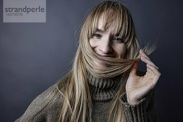 Junge Frau mit langem  blonden Haar hält lächelnd eine Haarsträhne vor den Mund  Porträt  Studioaufnahme