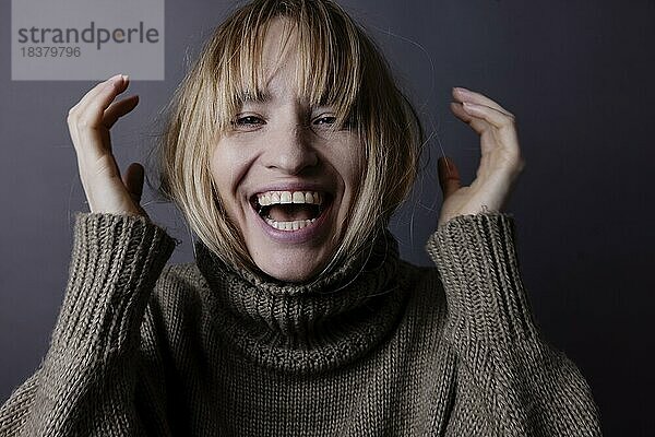 Junge Frau versteckt ihr langes  blondes Haar in einem Rollkragenpullover und lacht in Kamera  Portrait  Studioaufnahme