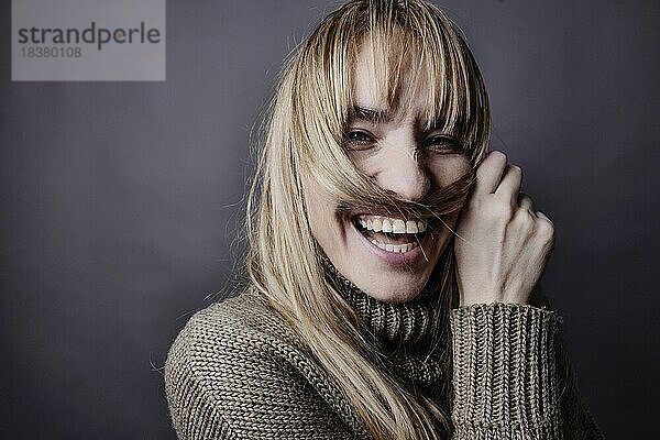 Junge Frau mit langem  blonden Haar macht sich aus einer Haarsträhne einen Schnurrbart und lächelt in Kamera  Portrait  Studioaufnahme