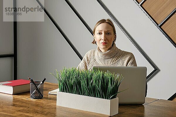 Selbstbewusste Frau arbeitet im Büro und schaut in die Kamera. Eine Topfpflanze steht auf ihrem Schreibtisch
