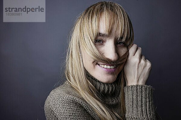 Junge Frau mit langem  blonden Haar macht sich aus einer Haarsträhne einen Schnurrbart und lächelt in Kamera  Portrait Studioaufnahme